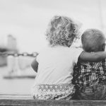 Schüßler Salze für Babys und Kleinkinder, hier zu sehen Geschwister von hinten, auf einer Bank sitzend, das Ältere legt den Arm um das Jüngere