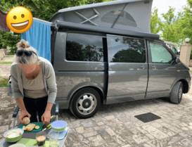 vor grauem VW Bus steht Frau in gebeugter Haltung und macht Essen